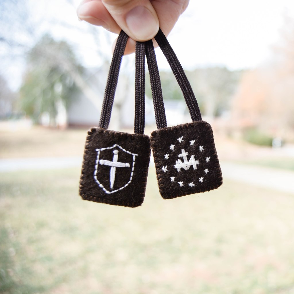 St. Michael Shield and Miraculous Medal back in white embroidery on brown scapular, held in front of blurry background