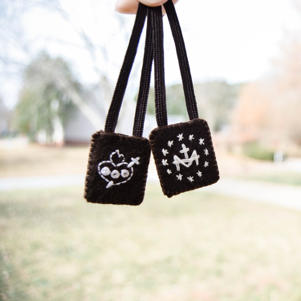Immaculate Heart of Mary and Miraculous Medal Back embroidered in white on brown scapular in front of blurry background